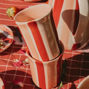 Può includere: Due tazze in ceramica impilate su una tovaglia a scacchi rossa e bianca. Le tazze presentano strisce verticali nei toni del rosso e del rosa. Sono visibili anche altri pezzi in ceramica coordinati, tra cui una brocca e una ciotola. La scena è decorata con petali di fiori e una fragola.