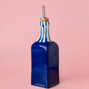 Op de afbeelding: Een blauw-wit gestreepte keramische fles met een roestvrijstalen tuit en een kurken stopper. De fles heeft een rechthoekige vorm en is geschilderd met een glanzende afwerking. De achtergrond is effen roze.