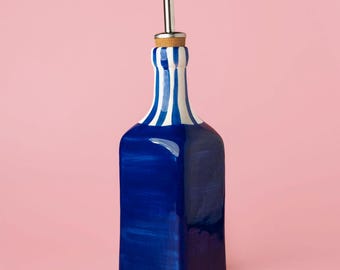 May include: A blue and white striped ceramic bottle with a stainless steel spout and cork stopper. The bottle has a rectangular shape and is painted with a glossy finish. The background is a solid pink colour.