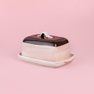 Puede incluir: Una mantequera de cerámica de forma rectangular. La base y los lados son de color rosa claro, mientras que la tapa es marrón oscuro con un asa blanca. El plato se encuentra sobre una bandeja rectangular a juego, todo sobre un fondo rosa.