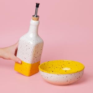 Puede incluir: Una botella de aceite de cerámica blanca y amarilla con una boquilla de acero inoxidable y un cuenco a juego. La botella y el cuenco tienen un diseño moteado. La parte inferior de la botella y el interior del cuenco son amarillos.