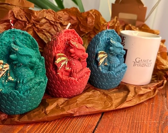 May include: Three dragon egg candles, one green, one red, and one blue, with gold accents. The candles are in the shape of dragon eggs with baby dragons inside. The candles are on a brown paper background.