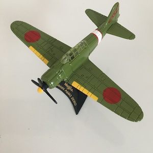 Puede incluir: Un modelo de avión verde con detalles rojos y amarillos, con una hélice y una cabina transparente. Las alas muestran círculos rojos. El modelo está montado sobre un soporte negro, con el texto "Maisto Mitsubishi A6M2 ZERO".