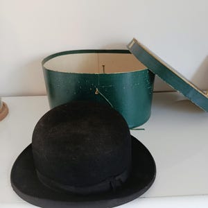 Op de afbeelding: Een zwarte bowlerhoed met een zwarte lintband ligt voor een groene hoedendoos met een gedeeltelijk open deksel. De hoed heeft een ronde kroon en een kleine rand. De hoedendoos is ovaal.
