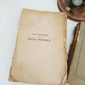 May include: An open, aged book with the title "TRAITE METHODIQUE DE MAGIE PRATIQUE" printed in black ink. The book's pages are yellowed and show signs of wear, with a torn corner. Another book is partially visible on the right.