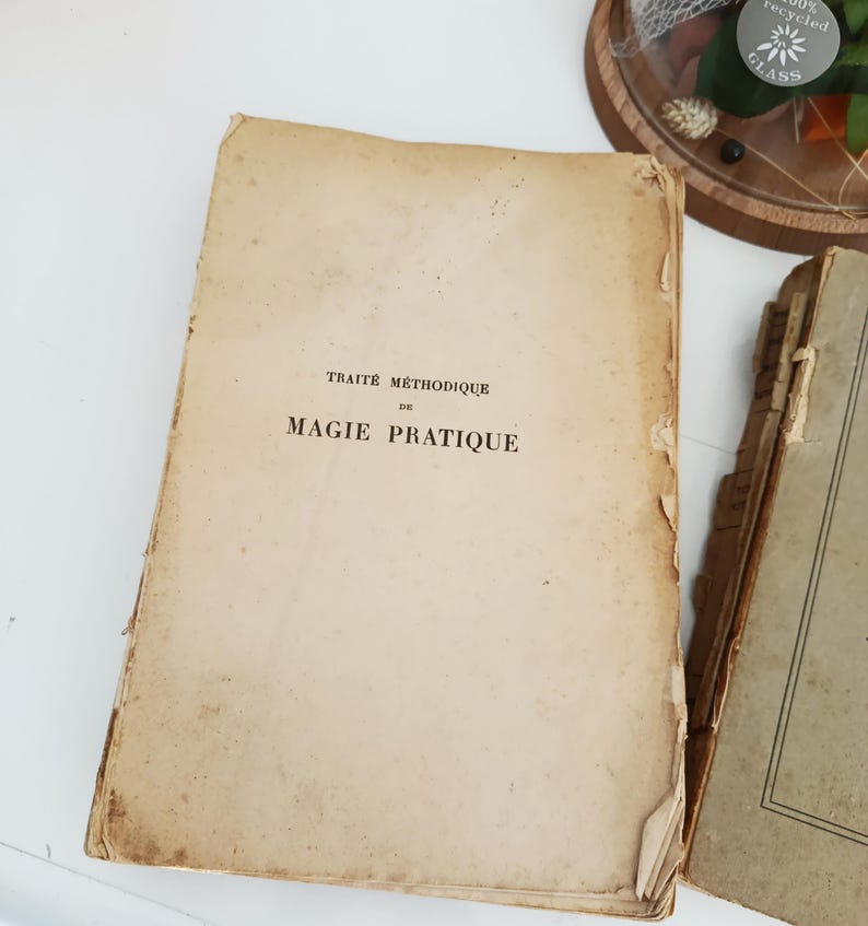 May include: An open, aged book with the title "TRAITE METHODIQUE DE MAGIE PRATIQUE" printed in black ink. The book's pages are yellowed and show signs of wear, with a torn corner. Another book is partially visible on the right.