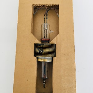 Puede incluir: Un tubo de vacío vintage en una caja de cartón. El tubo es de vidrio y tiene una base de metal. El tubo está rodeado por una bobina de metal.