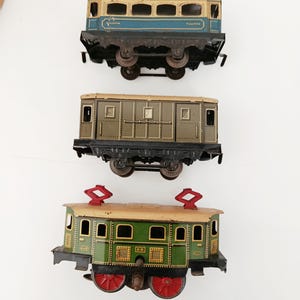 Peut inclure: Trois wagons de train jouets en fer-blanc vintage disposés verticalement. Le wagon supérieur est bleu et crème avec le texte Pullman. Le wagon central est vert olive avec un design à panneaux. Le wagon inférieur est vert avec des roues rouges.