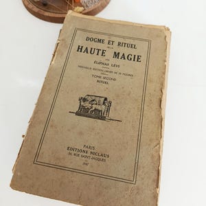 May include: A vintage book titled "DOGME ET RITUEL DE LA HAUTE MAGIE" by &Eacute;liphas L&eacute;vi. The cover is a faded tan color with black text and illustrations. The book is a second edition, published in Paris in 1947.