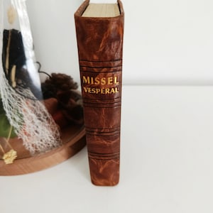 May include: A vintage book with a brown leather cover. The title "MISSEL VESPERAL" is embossed in gold lettering. The book's spine features horizontal embossed lines. The pages are aged, and the book is standing upright.
