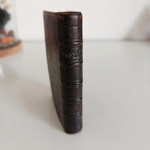Può includere: Un piccolo libro antico con una copertina in pelle marrone scuro e strutturata. Il dorso presenta fasce in rilievo e testo in rilievo. Il libro è in posizione verticale, mostrando il suo aspetto invecchiato e il suo design classico. I bordi del libro sono leggermente usurati.