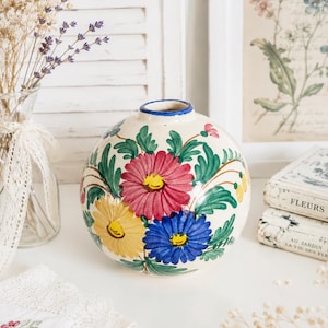 May include: A round ceramic vase with a cream-colored base featuring hand-painted floral motifs in red, yellow, and blue with green leaves. The vase has a narrow, circular opening with a thin blue rim, showcasing a smooth, glossy finish.