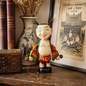 Op de afbeelding: Een vintage-stijl beeldje van een lachend personage met een kaal hoofd, twee plukjes donker haar en rode oorbellen. Het figuur draagt een metallic oranje en groen gewaad met rode accenten, staat op een zwarte voet en houdt kleine metalen bellen vast.