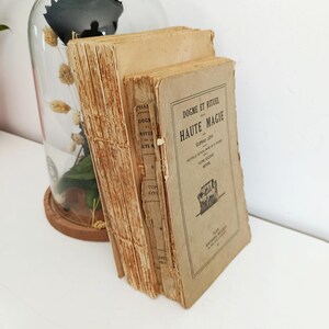 May include: A stack of antique books with aged, tan covers and deckled edges. The book on the right has the title "HAUTE MAGIE" printed in black. A glass cloche with dried flowers and a dark object is in the background.