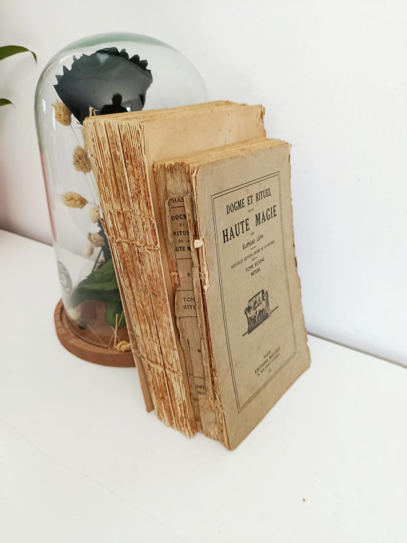 May include: A stack of antique books with aged, tan covers and deckled edges. The book on the right has the title "HAUTE MAGIE" printed in black. A glass cloche with dried flowers and a dark object is in the background.