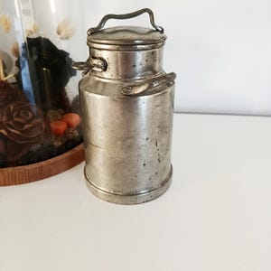 May include: An antique metal milk churn with a handle on top and side handles. The churn is cylindrical with a lid and is made of a silver-coloured metal. The churn is on a white surface.