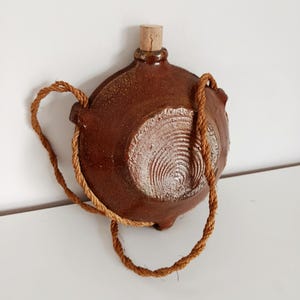 Könnte beinhalten: Eine braune Keramikflasche mit Korken und Seilgriff. Die Flasche hat ein kreisförmiges Design auf der Vorderseite und wird wahrscheinlich zum Tragen von Flüssigkeiten verwendet. Der Seilgriff ist an den Seiten der Flasche befestigt.
