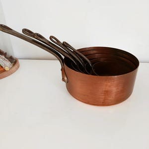 May include: A set of copper saucepans with long, dark metal handles. The saucepans are nested inside each other, showcasing their varying sizes. The copper has a warm, reddish-brown hue, and the handles are riveted to the sides.