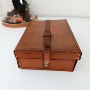 Könnte beinhalten: Eine rechteckige, braune Lederbox mit Riemenverschluss. Die Box hat ein leicht abgenutztes Aussehen mit sichtbaren Nähten und Metallnieten. Der Riemen ist geschnallt und mit zwei Metallknöpfen befestigt. Die Box wird wahrscheinlich zur Aufbewahrung verwendet.