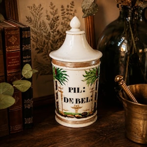 Op de afbeelding: Een witte keramische apothekerspot met bijpassend deksel, voorzien van een handgeschilderd ontwerp van groene palmbomen en een centraal caduceussymbool. De pot toont de zwarte tekst PIL: DE BEL: en heeft bruin geschilderde randen aan de boven- en onderkant.