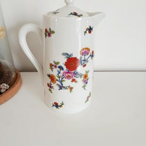 Peut inclure: Pichet en céramique blanche avec couvercle, orné de motifs floraux colorés. Le pichet a une anse incurvée et un bec verseur. Le motif floral comprend des fleurs rouges, roses, oranges et bleues. Un objet décoratif pour servir des boissons.