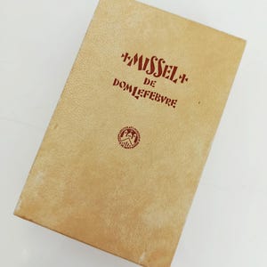 Pode incluir: Uma caixa retangular castanha clara com as palavras "MISSEL" e "DE DOM LEFEBVRE" impressas a vermelho. Um emblema circular é visível na parte inferior. A caixa tem uma superfície texturizada e parece ser feita de cartão.