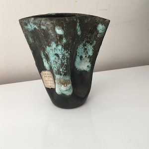May include: A dark green and blue ceramic vase with a unique, irregular shape. The vase has a label that reads "Eaux Neige de Savoie Fait Main Cuisiniere".
