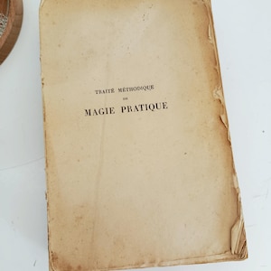 May include: An antique book with a worn, tan cover. The title "TRAITE METHODIQUE DE MAGIE PRATIQUE" is printed in black ink. The edges of the book show signs of age and wear, with some peeling.