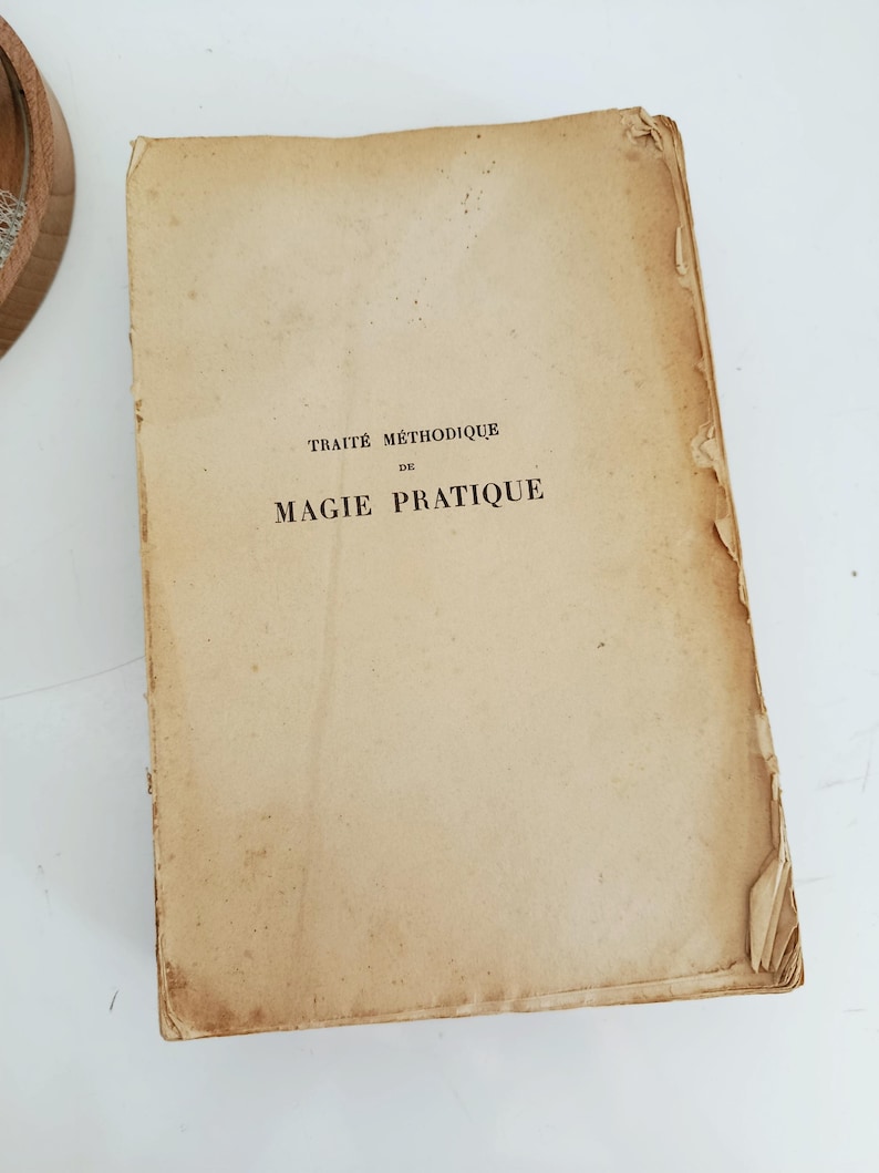 May include: An antique book with a worn, tan cover. The title "TRAITE METHODIQUE DE MAGIE PRATIQUE" is printed in black ink. The edges of the book show signs of age and wear, with some peeling.