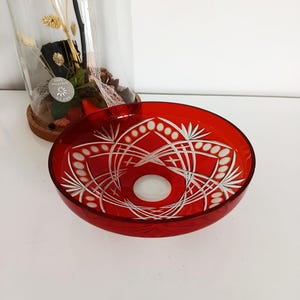 May include: A vibrant red glass bowl with intricate white etched floral patterns. The bowl has a circular shape and a smooth, glossy finish. The design features a central circle and radiating lines. The bowl is on a white surface.