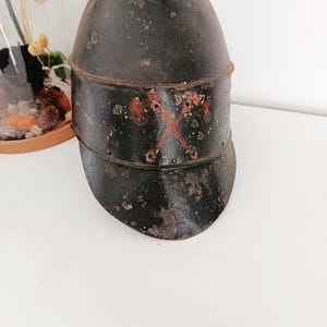 Peut inclure: Casque en métal noir vieilli avec visière. Le casque présente des signes d'usure, avec de la rouille et des marques rouges. Il comporte un bord et une bande au milieu. Il s'agit probablement d'un objet historique.