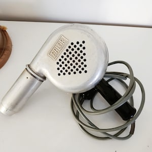 May include: A vintage silver-coloured hair dryer with the word "CALOR" printed on the front. The dryer has a black handle and a grey cord. The head of the dryer has a series of small holes.