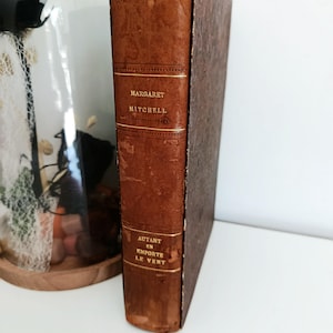 Peut inclure: Un livre ancien à couverture rigide en cuir marron usé. Le dos porte le nom de l'auteur, Margaret Mitchell, et le titre "Autant en emporte le vent" en lettres dorées. Le livre est présenté sur une surface blanche.