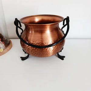 Op de afbeelding: Een koperen ketel met een gehamerde textuur, voorzien van zwarte metalen handvatten en drie poten. De ketel heeft een glanzend, reflecterend oppervlak en is ontworpen voor decoratieve doeleinden.