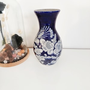 Peut inclure: Un vase en céramique bleu foncé avec un motif floral blanc. Le vase a un dessus évasé et un corps arrondi. Le motif floral est détaillé et couvre la majeure partie de la surface du vase. C'est un objet décoratif.