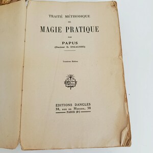 May include: An open book with the title "TRAIT&Eacute; M&Eacute;THODIQUE DE MAGIE PRATIQUE" by Papus. The book is an antique with aged, off-white pages and black text. The cover includes the publisher's name and address in Paris.
