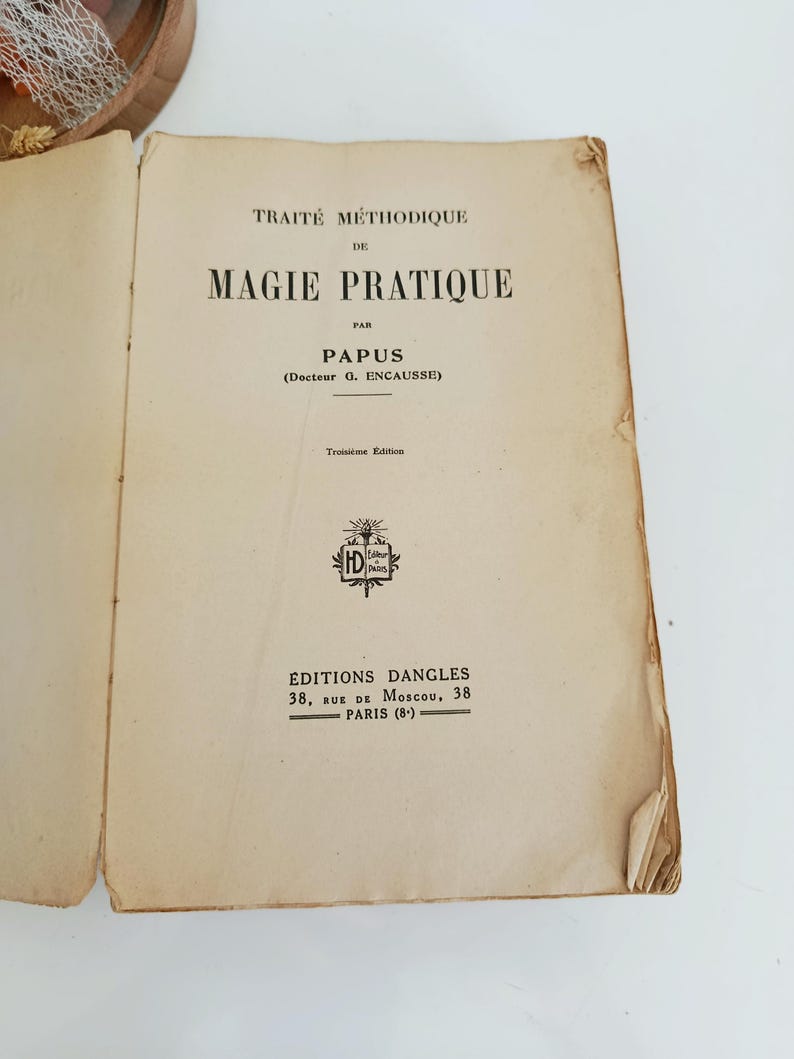 May include: An open book with the title "TRAIT&Eacute; M&Eacute;THODIQUE DE MAGIE PRATIQUE" by Papus. The book is an antique with aged, off-white pages and black text. The cover includes the publisher's name and address in Paris.