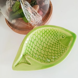 Pode incluir: Um prato de cerâmica verde claro em forma de peixe com um padrão de escamas texturizadas. O prato tem um design de barbatana caudal e um pequeno detalhe de olho. Uma cúpula de vidro com plantas secas e elementos decorativos está no fundo.