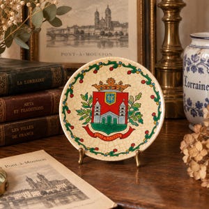 Op de afbeelding: Een ronde decoratieve keramische schaal met een crèmekleurige craquelé glazuurafwerking. Het midden toont een kleurrijk wapenschild met een rood schild, twee torens en een kroon. Een groen-rode bloemenrand omringt het embleem met de tekst PONT-A-MOUSSON.