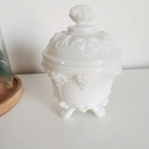 Puede incluir: Un tarro blanco con tapa de estilo vintage con un patrón decorativo de uvas y hojas. El tarro tiene tres patas pequeñas y un asa detallada en la tapa. El diseño general es ornamentado y elegante, adecuado para exhibición o almacenamiento.