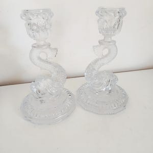 May include: A pair of clear glass candle holders. Each features a dolphin-shaped stem, a round base, and a cup-shaped top. The glass has a textured finish, and the candle holders are designed to hold candles.