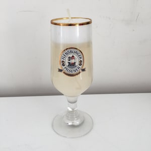May include: A clear glass candle shaped like a beer glass with a gold rim. The candle is filled with a white wax and has a gold label with the text "Flensburger Pilsener" and a ship logo.