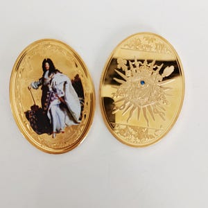 May include: Two gold-coloured oval coins. One coin features a portrait of a person in historical attire. The other coin has a sunburst design with a crown and a blue gem in the centre. Both coins have ornate borders.