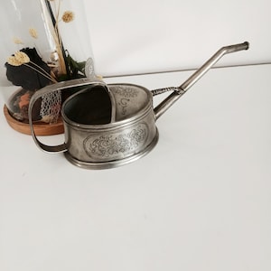 May include: An antique silver watering can with an ornate floral design. The can has a long spout and a handle. The design is etched into the metal, giving it a vintage look. It is likely used for watering small plants or flowers, and is a decorative item.