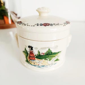 Peut inclure: Pot en céramique crème avec couvercle, orné d'une bordure florale. Le pot présente une scène peinte d'une femme près d'oies et d'une maison. Le pot a deux petites poignées.
