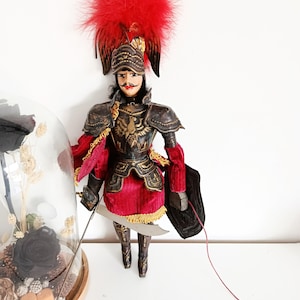 Puede incluir: Muñeca decorativa vestida de soldado, con un sombrero de plumas rojas y una armadura detallada. La muñeca sostiene una espada y un escudo, con una falda de terciopelo rojo y adornos dorados. La muñeca mide unos 30 cm de altura.