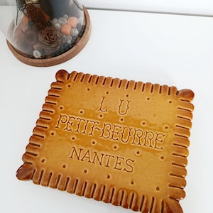 Peut inclure: Un plat de service en céramique en forme de biscuit, de couleur brun doré. Le plat porte l'inscription en relief "L.U. PETIT-BEURRE NANTES" et des détails décoratifs sur les bords. Le plat est rectangulaire avec des coins arrondis.