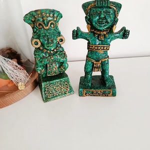 Peut inclure: Deux statues vertes et dorées de figures anciennes. Les statues présentent des détails complexes et reposent sur des socles rectangulaires. Les figures portent des coiffes et des bijoux. L'une des statues a les bras tendus. Les statues sont sur une surface blanche.