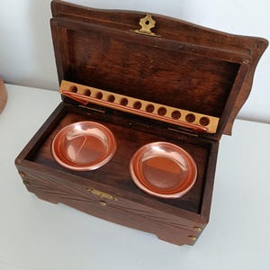 May include: Open wooden box with two copper-colored bowls and a row of holes. The box is dark brown with carved details and a brass clasp. The lid is open, revealing the interior. The box is on a white surface.