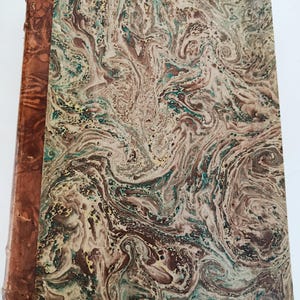 May include: Antique book with a marbled cover in shades of brown, cream, and teal. The spine is a darker brown leather. The cover has a swirling pattern, giving it a unique and vintage appearance. The book is likely a collectible or decorative item.